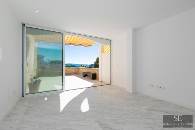 Bright empty room with white marble floors and a large sliding glass door opening to a sunny stone terrace.