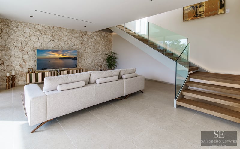 Spacious living room featuring a beige sofa, a decorative stone wall with a TV, and wooden stairs with a glass railing.