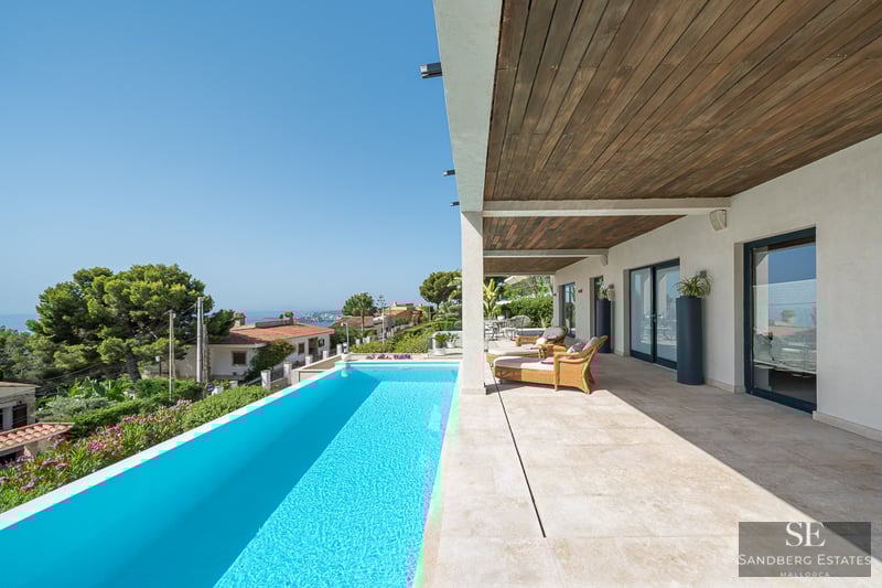 Sonnige Terrasse mit türkisfarbenem Infinity-Pool, Korbliegen und Holzdecke mit Blick auf die Küste.