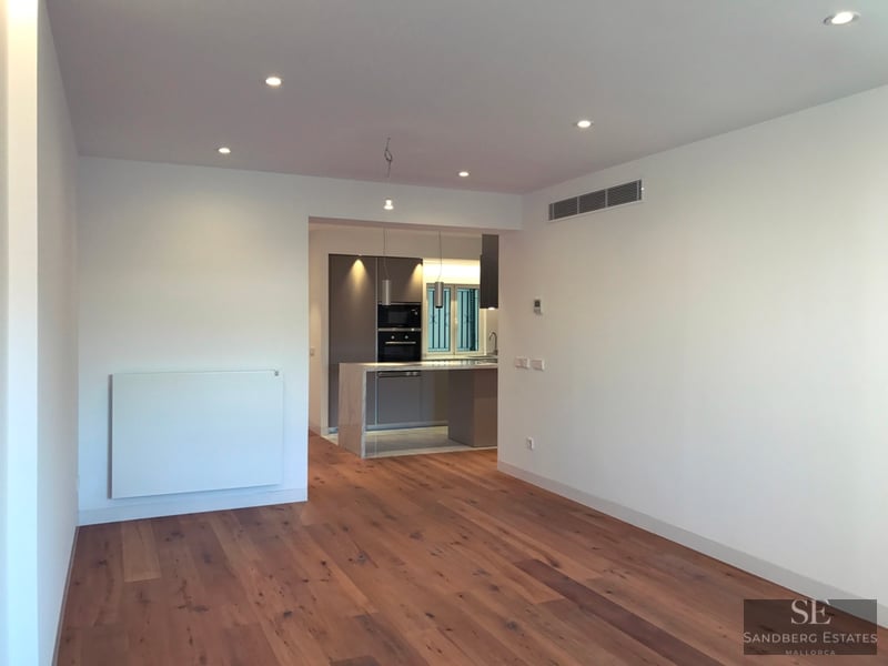 Spacious empty living room with hardwood floors leading to a modern grey kitchen with recessed ceiling lights.