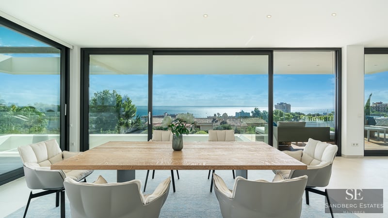 Bright dining room with a large wooden table and grey chairs facing floor-to-ceiling windows with a vast ocean view.