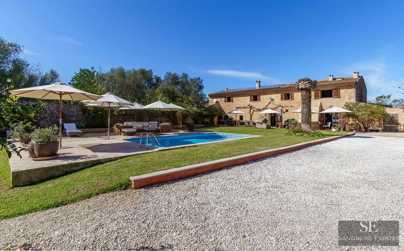 View of a large stone villa with a blue swimming pool, white umbrellas, and garden under a clear sky.