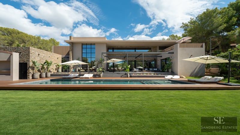 Contemporary villa featuring a lap pool, wood decking, green lawn, and stone facade under a bright blue sky.