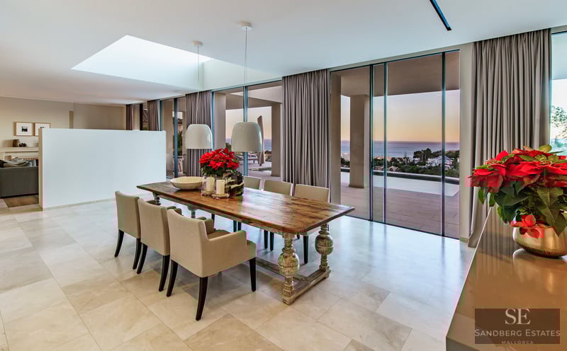 Elegant dining area featuring a long wooden table and floor-to-ceiling windows overlooking the ocean at sunset.