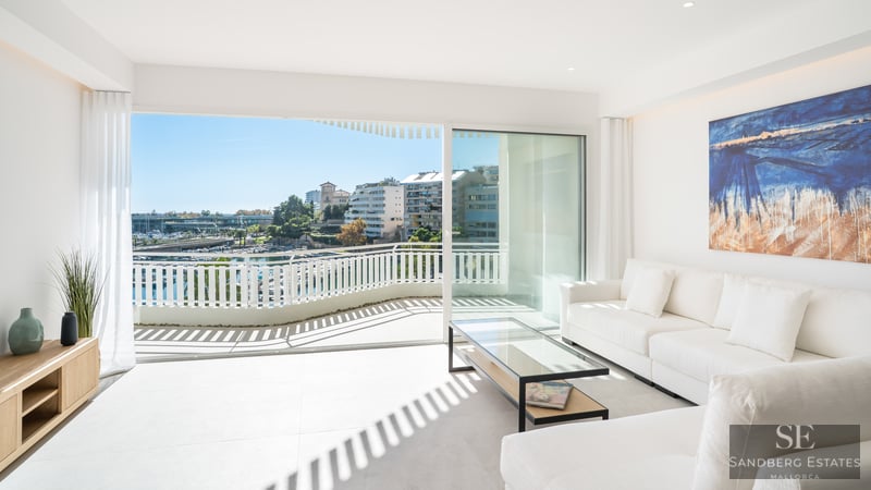 Bright living room with white sofas and glass doors opening to a balcony overlooking a marina.