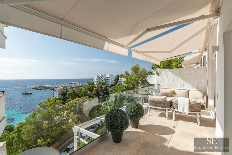 Modern terrass med beige soffor, glasräcke och panoramautsikt över havet under markiser.