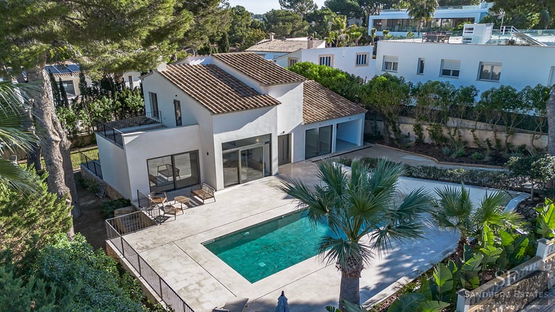 Aerial view of a white villa with a tiled roof, turquoise swimming pool, and large stone terrace surrounded by pine and palm trees.
