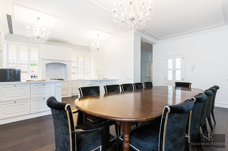 Large oval dark wood dining table surrounded by black velvet chairs under ornate crystal chandeliers in a white room.