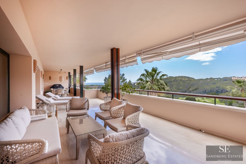 Grand balcon couvert avec mobilier en osier offrant une vue sur une vallée verdoyante et la mer par temps clair.