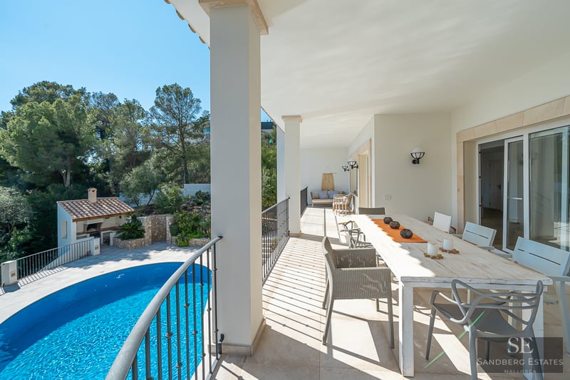 Terrasse couverte blanche avec grande table à manger et vue sur une piscine bleue et de la verdure.
