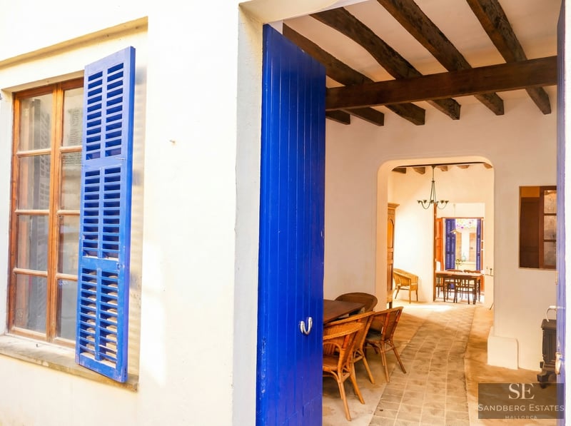 White exterior wall with bright blue shutters and open door leading to a rustic interior with wooden beams.