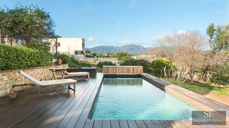 A rectangular swimming pool with a dark wood deck, white sun loungers, and a scenic mountain backdrop.