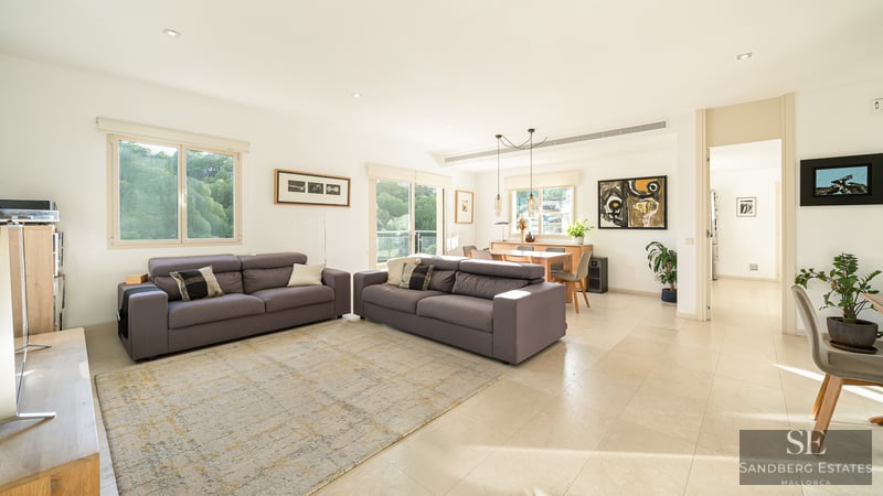 Spacious bright living room with two grey sofas, light tile flooring, and dining area in the background with large windows.