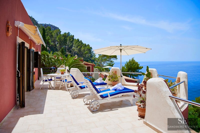 Solig terrass med blå solstolar, vitt parasoll och panoramautsikt över Medelhavet och klipporna.