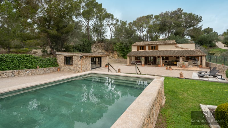 Piscina exterior con bordes de piedra frente a una villa rústica mediterránea rodeada de pinos.