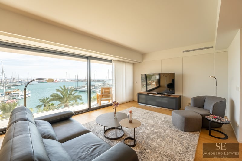 Bright modern living room with a black leather sofa and large windows overlooking a sunny marina with boats.