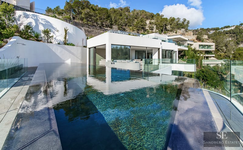 Modern infinity pool with clear blue water overlooking a white minimalist villa nestled in a green hillside.