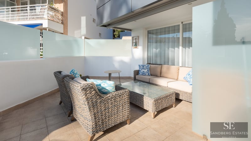 Modern terrass med grå soffor i konstrotting, beige dynor och blå kuddar på en solig stenlagd uteplats.