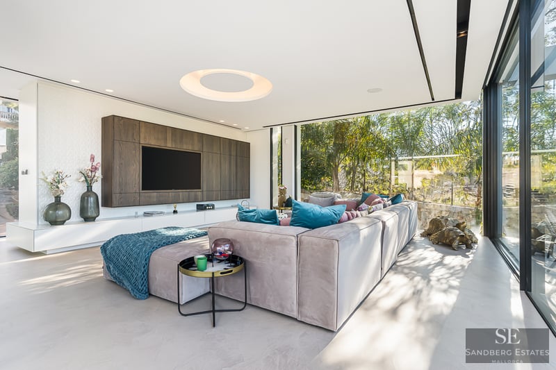 A bright contemporary living room with a large sectional sofa, wall-mounted TV, and floor-to-ceiling garden windows.