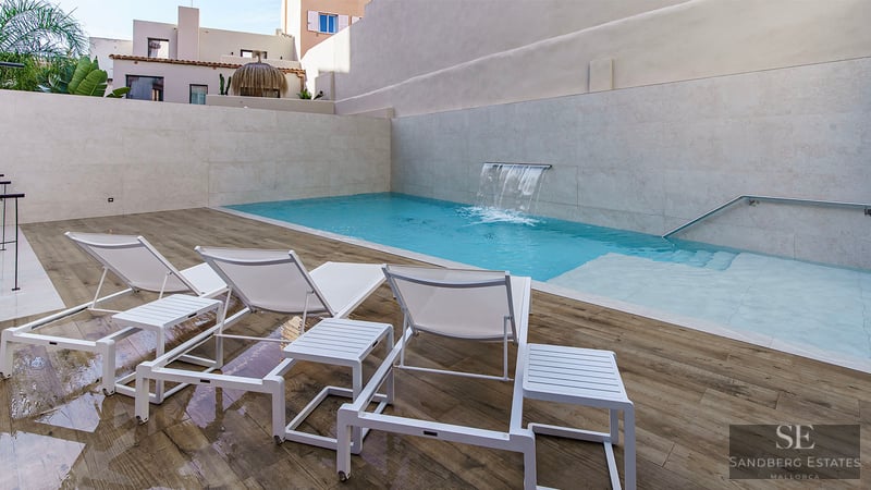 Türkisfarbener Pool mit integriertem Wasserfall, weißen Liegen auf Holzdeck und Steinmauern.