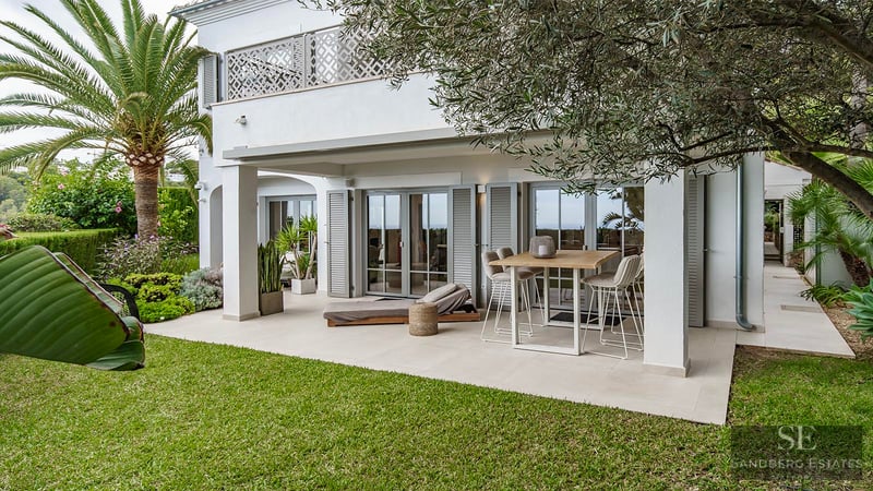 Villa mediterránea blanca con porche cubierto, mesa alta, tumbona, césped verde y palmeras.