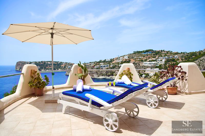 Två blå solstolar och ett vitt parasoll på en stenterrass med panoramautsikt över havet och kusten.