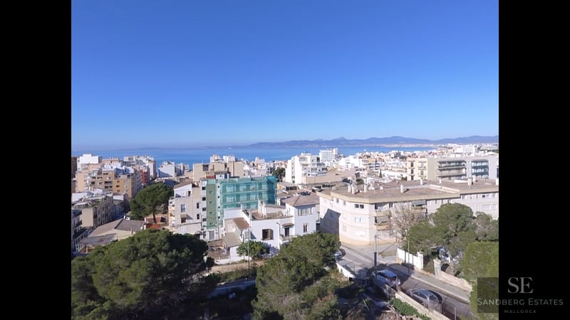 Elevated view of a Mediterranean coastal city with white buildings, green trees, and the blue sea on the horizon.
