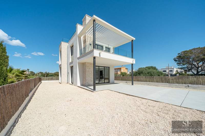 Two-story white modern villa with natural stone walls, a large balcony, and a gravel courtyard under a clear blue sky.