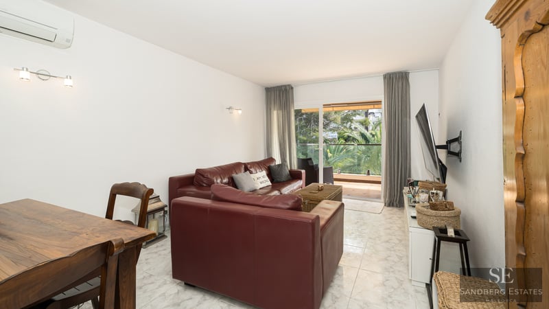 Modern living room featuring burgundy leather sofas, white marble floors, and a glass door view to a lush green balcony.
