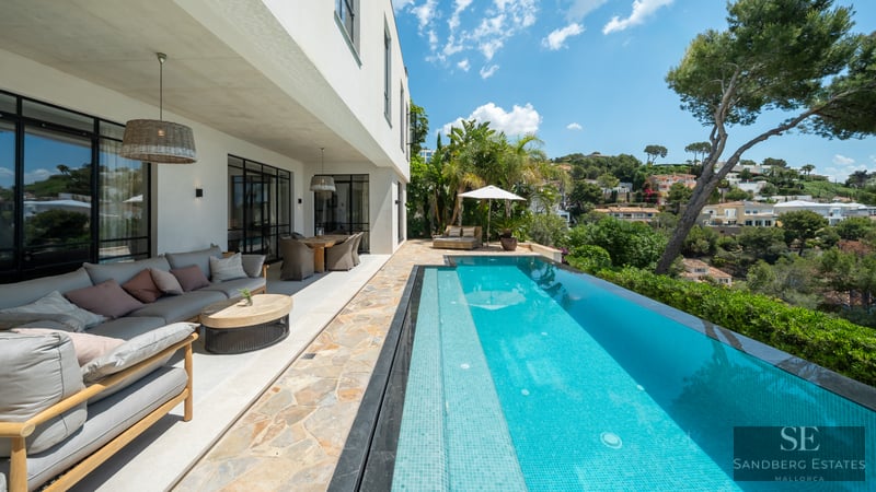 Luxury outdoor terrace with a long turquoise pool, grey sofa lounge, dining area, and views of a green hillside.