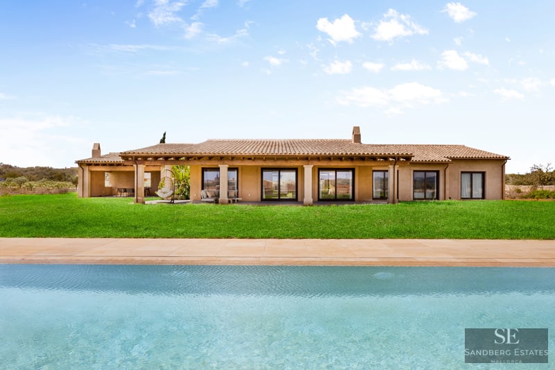 Large turquoise pool in front of a modern Mediterranean villa with a green lawn and terracotta roof under a blue sky.
