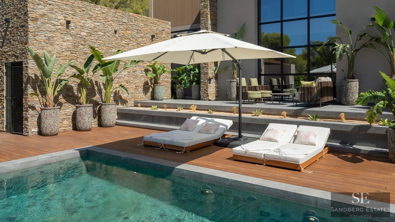 Modern pool area with wooden deck, white sun loungers, and large umbrella against a natural stone wall.