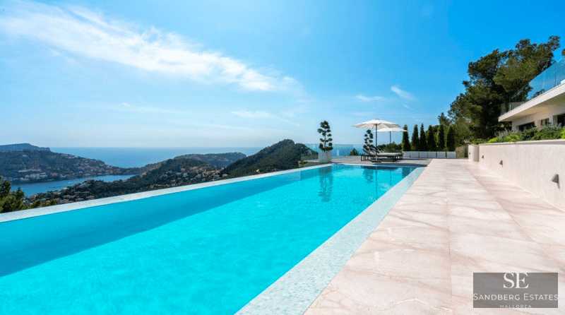 Turquoise infinity pool overlooking the sea and mountains, surrounded by light stone tiles and white umbrellas.