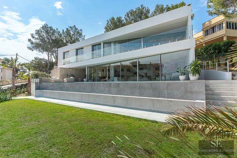 White modern villa with large windows, infinity pool, and green lawn under a sunny sky.