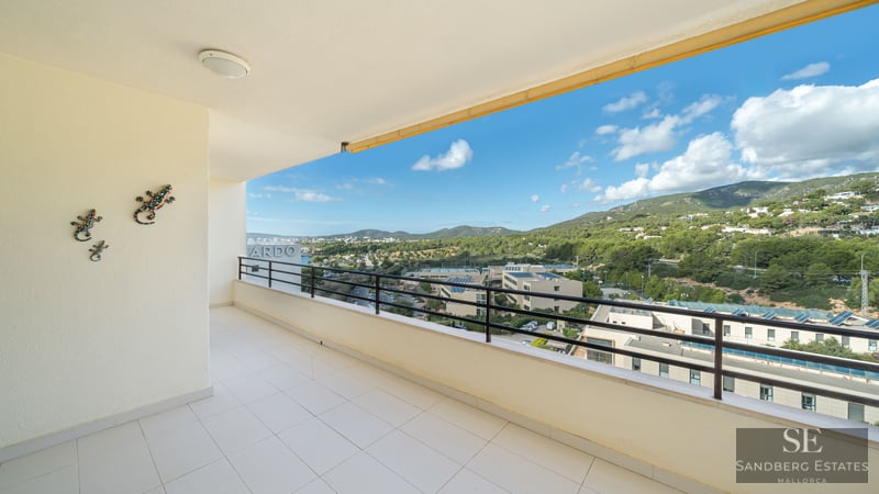 Spacious white tiled balcony with a black metal railing overlooking green hills and a blue sky with white clouds.
