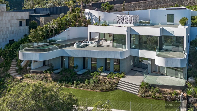 High-angle view of a multi-level white modern villa with an infinity pool, curved terraces, and lush gardens.