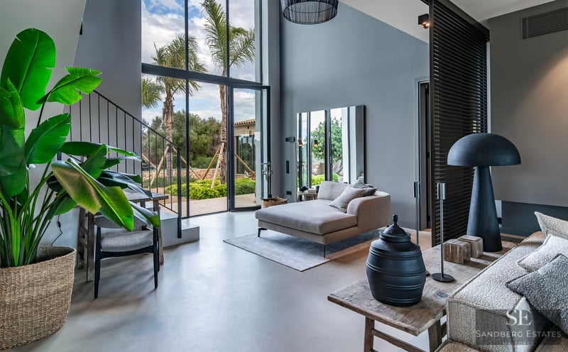 Spacious living area with high ceilings, floor-to-ceiling windows with palm tree views, and modern grey chaise lounge.