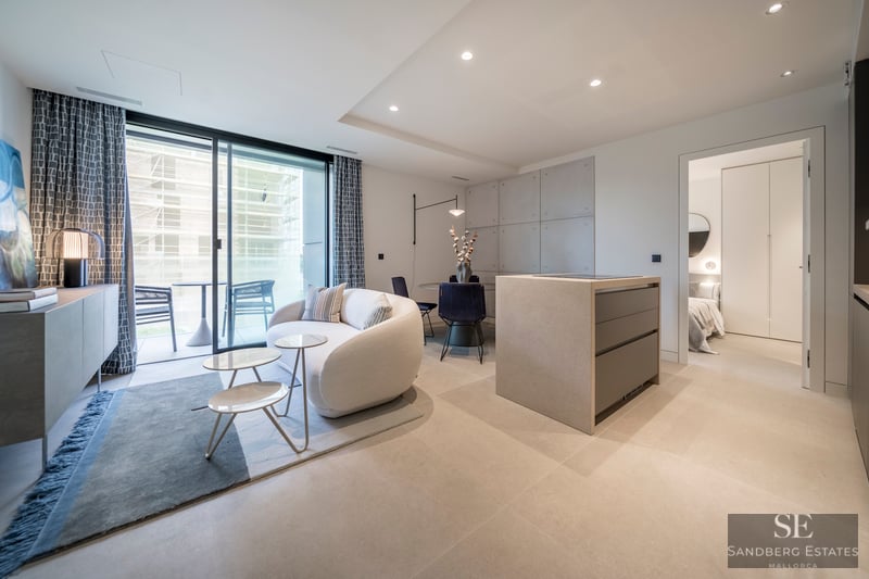 Sala de estar moderna con sofá curvo blanco, isla de cocina de piedra y grandes puertas de cristal hacia una terraza.
