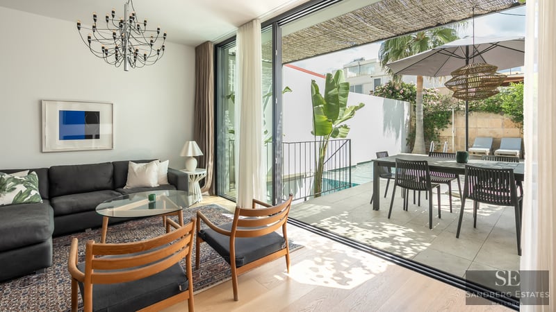 Salón luminoso con sofá oscuro y sillas de madera que se abre a una terraza soleada con piscina y comedor.