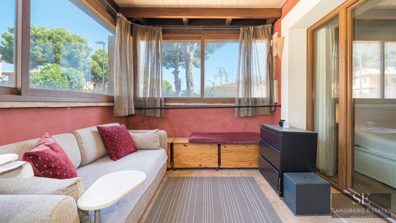 Sunlit living area with beige sofa, wooden beams, terracotta walls, and large windows overlooking green trees.