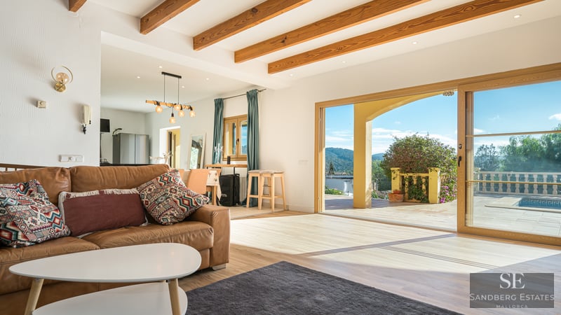 Living room with brown leather sofa, exposed wood beams, and large sliding doors opening to a terrace with mountain views.