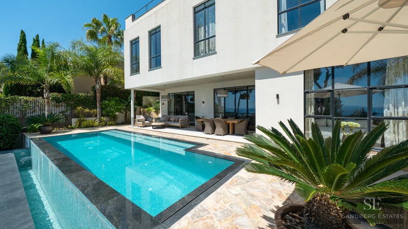 Bright outdoor scene featuring a turquoise infinity pool, covered terrace, and modern white villa with palm trees.