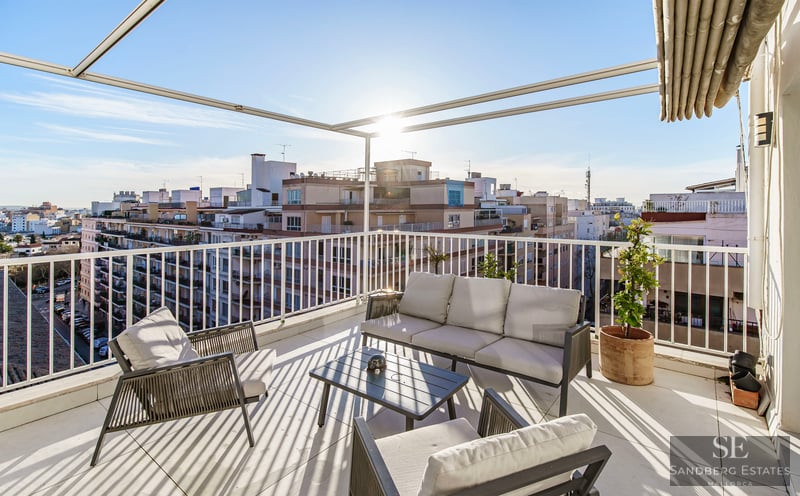 Sun-drenched rooftop terrace with modern grey outdoor furniture, white railings, and an expansive city skyline view.
