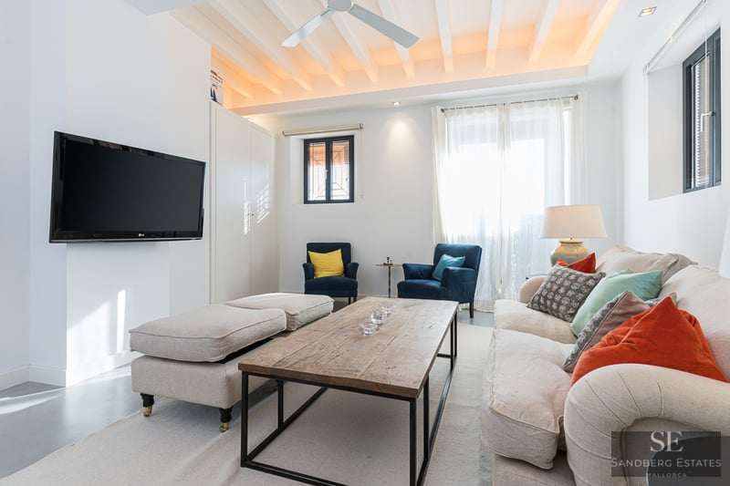 Ljust vardagsrum med vitt bjälktak, beige soffa med färgglada kuddar och väggmonterad TV.