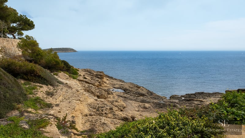 Rocky coastline meeting the blue sea with green Mediterranean shrubs in the foreground.