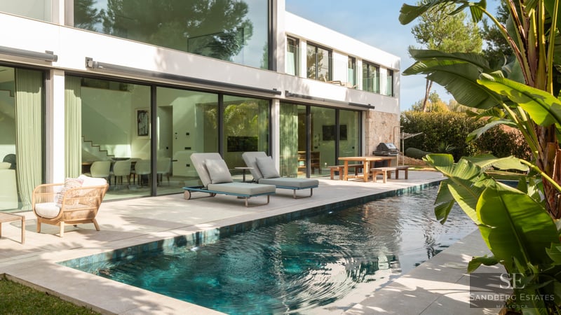 Turkost pool bredvid en modern vit villa med stora glasfönster och solsängar.