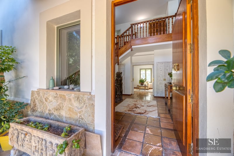 Puerta de madera abierta que da a un recibidor mediterráneo con suelos de terracota y escalera de madera.