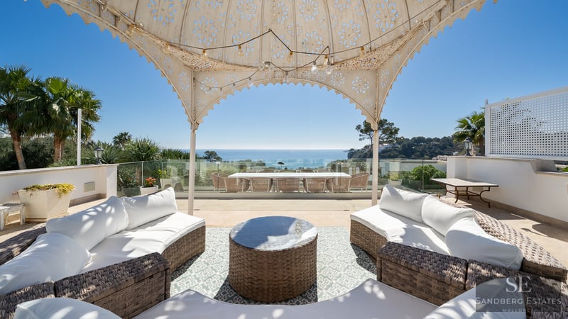 Amplia terraza con sofás de mimbre bajo un cenador decorativo y vistas despejadas al océano Mediterráneo.