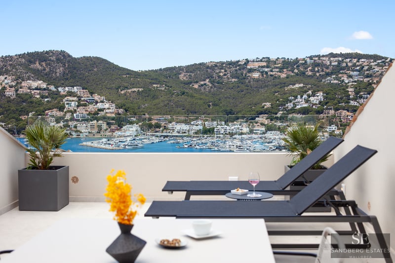 Sun loungers on a modern terrace overlooking a harbor with white boats and lush green hills under a clear blue sky.