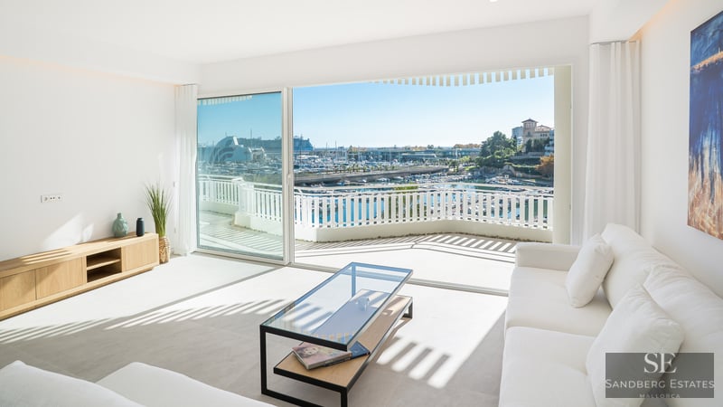 Bright minimalist living room with white sofas and large glass doors opening to a harbor view and terrace.